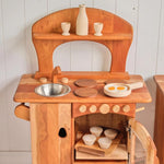 Wooden play kitchen accessory set shown on counter with milk jug, cups, and pans