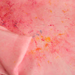 A close-up of a light pink fabric or paper material with scattered multicolored speckles and a folded corner showing the texture and color.