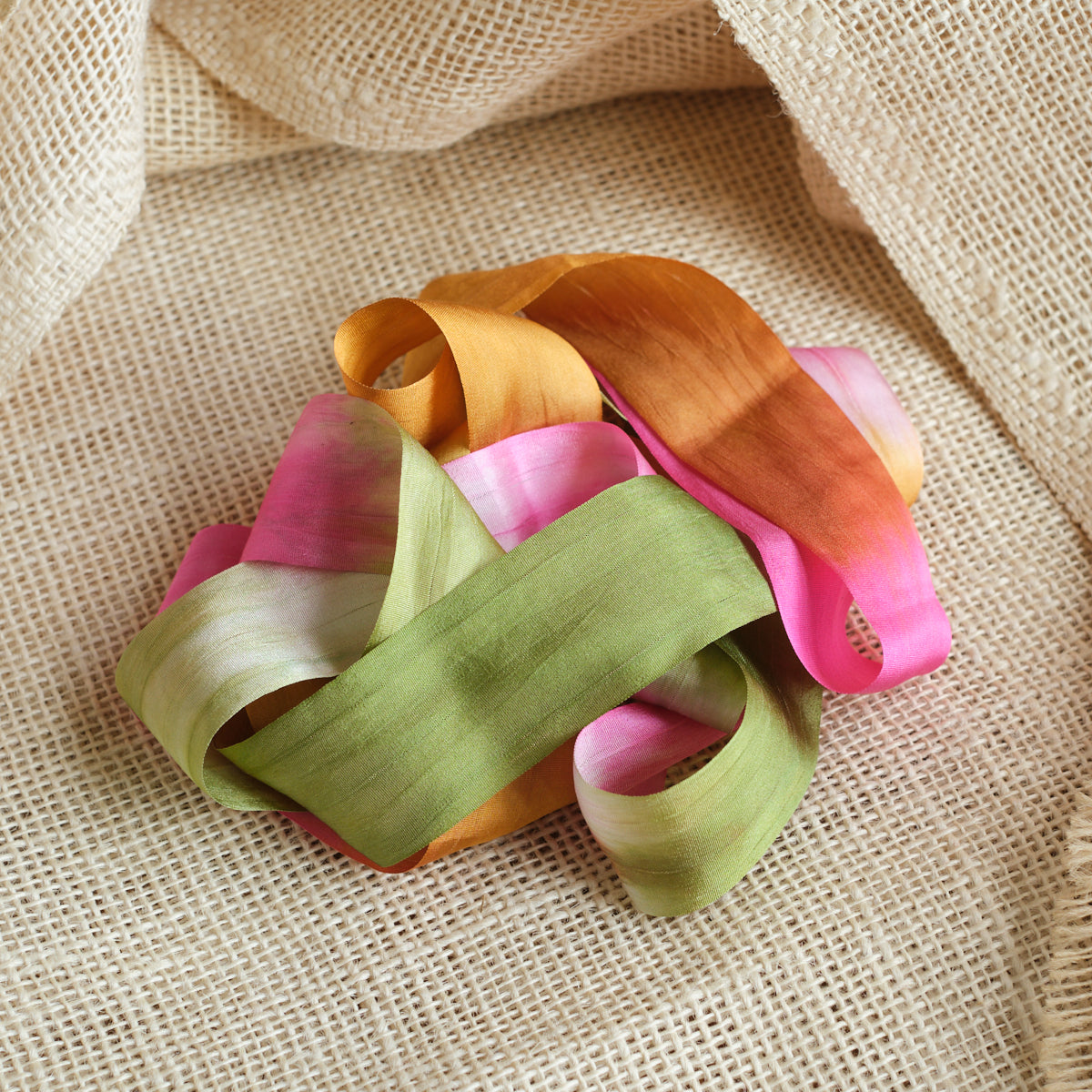 Green, pink, and golden brown ribbon artistically laying in a twisted fashion on a light brown textured fabric background.