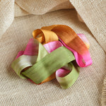 Green, pink, and golden brown ribbon artistically laying in a twisted fashion on a light brown textured fabric background.