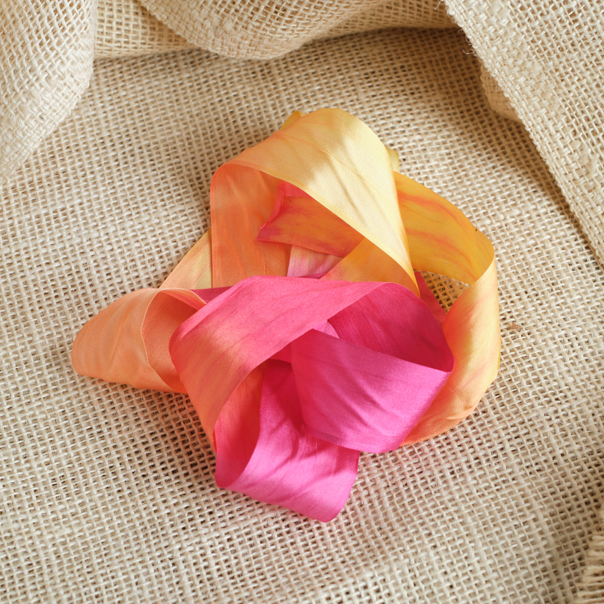Pink, Orange, and yellow ribbon artistically laying in a twisted fashion on a light brown textured fabric background.