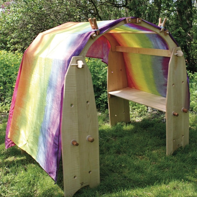 A small wooden play hide or tunnel with a rainbow-colored fabric lining, set outdoors on grass with trees in the background.